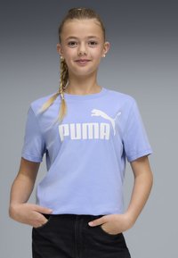 Νεαρή κοπέλα με ξανθό πλεξούδα, φοράει ένα ανοιχτό μωβ μπλουζάκι Puma και μαύρο παντελόνι, στέκεται με τα χέρια στις τσέπες μπροστά σε γκρι φόντο.