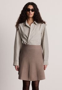 Camicia a righe beige e blu con bottoni, abbinata a una gonna marrone aderente. La modella indossa occhiali da sole neri e ha capelli lunghi e ricci.