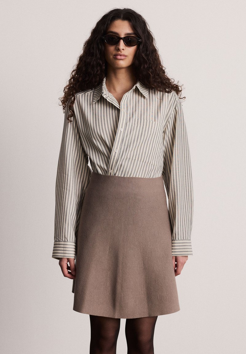 Camicia a righe beige e blu con bottoni, abbinata a una gonna marrone aderente. La modella indossa occhiali da sole neri e ha capelli lunghi e ricci.