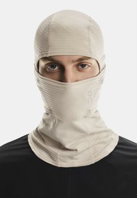 Person wearing a beige, full-face balaclava with visible eyes, paired with a black top, against a plain white background.