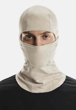Oseba, oblečena v bež balaklavo, ki pokriva celoten obraz in ima vidne oči, v kombinaciji s črno majico, na eni barvni beli podlagi.