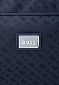 Marinefarget stoffpose har et teksturert mønster; senteretiketten viser "BOSS" i hvitt på en kontrastfargebakgrunn. Detaljene er minimale.