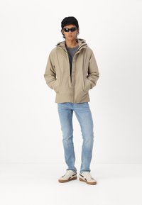 Beige waterdichte jas met capuchon, voorzakken, gedragen over een donkere blouse, gecombineerd met lichtblauwe jeans en witte sneakers. Zwarte zonnebril.