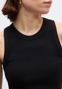 Κοντινό πλάνο γυναίκας που φοράει μαύρο, ριμπ, αμάνικο τοπ και χρυσό κρίκο σκουλαρίκι, στραμμένη προς τα δεξιά, μπροστά σε ουδέτερο φόντο.