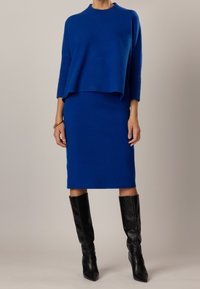 Pull tricot bleu à col montant et manches trois-quarts, assorti à une jupe bleue. Porté avec des bottes noires montantes jusqu'aux genoux.