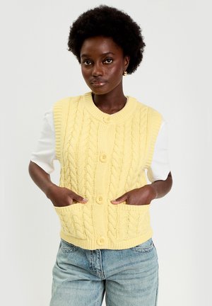 Giovane donna nera con capelli corti e ricci che indossa un gilet giallo lavorato a trecce con bottoni sopra una camicia bianca e jeans celesti, con le mani nelle tasche.