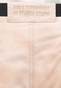 Tissu beige avec une couture verticale et une étiquette cousue portant l'inscription "AIGLE EXPERIENCE BY ÉTUDES STUDIO" attachée à une sangle noire.