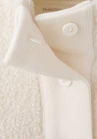Gros plan sur un poignet en tissu côtelé couleur crème avec deux boutons blancs sur un col de veste crème texturé et duveteux.