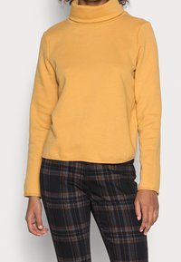 Jersey de cuello alto amarillo en una tela suave y elástica, combinado con pantalones de cuadros negros con líneas rojas y azules. Textura suave.