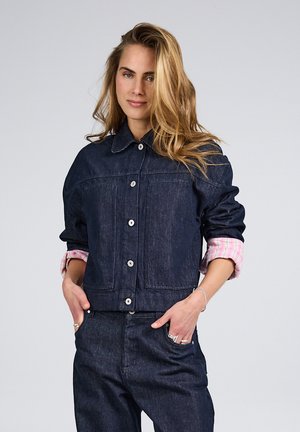 Frau mit langen blonden Haaren, die eine dunkle Jeansjacke und Jeans trägt, die Hände in den Taschen, vor einem schlichten hellen Hintergrund.