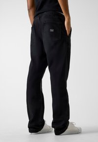 Pantalon en denim noir à coupe décontractée, avec une taille élastique, deux poches arrière et une discrète étiquette logo sur la poche droite.