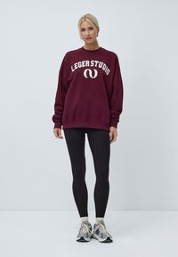 Bordeaux genser med "LEGERSTUDIO" logo, løs passform, kombinert med svarte leggings og grå joggesko. Ensfarget bakgrunn; modell som står.
