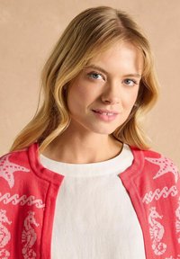 Růžový kardigan s bílými vzory mořského koníka a vln, vyrobený z měkkého pleteného materiálu. Nosený přes jednoduché bílé tričko s kulatým výstřihem.