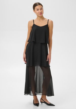 Une femme porte une robe noire à fines bretelles et à plusieurs épaisseurs, avec une jupe transparente à pois et des talons noirs, sur un fond uni.