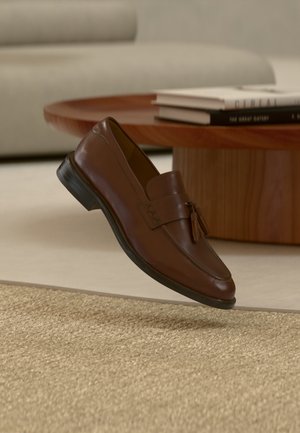Mocassin à pampilles en cuir marron flottant au-dessus d'un tapis beige texturé, avec une table ronde en bois et des piles de livres en arrière-plan.