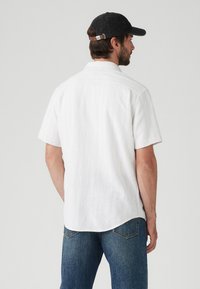 Camicetta bianca a maniche corte con righe verticali, orlo curvo e vestibilità rilassata, abbinata a jeans blu e un cappellino da baseball scuro.