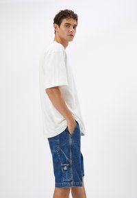 Camiseta blanca de manga corta con un corte relajado, combinada con pantalones cortos de mezclilla azul con bolsillos y detalles de costura. Textura de tela suave.