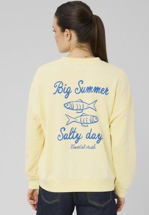 Femme aux cheveux bruns attachés en queue de cheval, portant un sweat-shirt jaune pâle avec un texte bleu et des illustrations de poissons au dos.