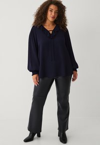 Marineblauwe blouse met ruches bij de hals en strikdetail, gecombineerd met zwarte neplederen broek. Beide kledingstukken hebben een losse pasvorm en een textiele stof.