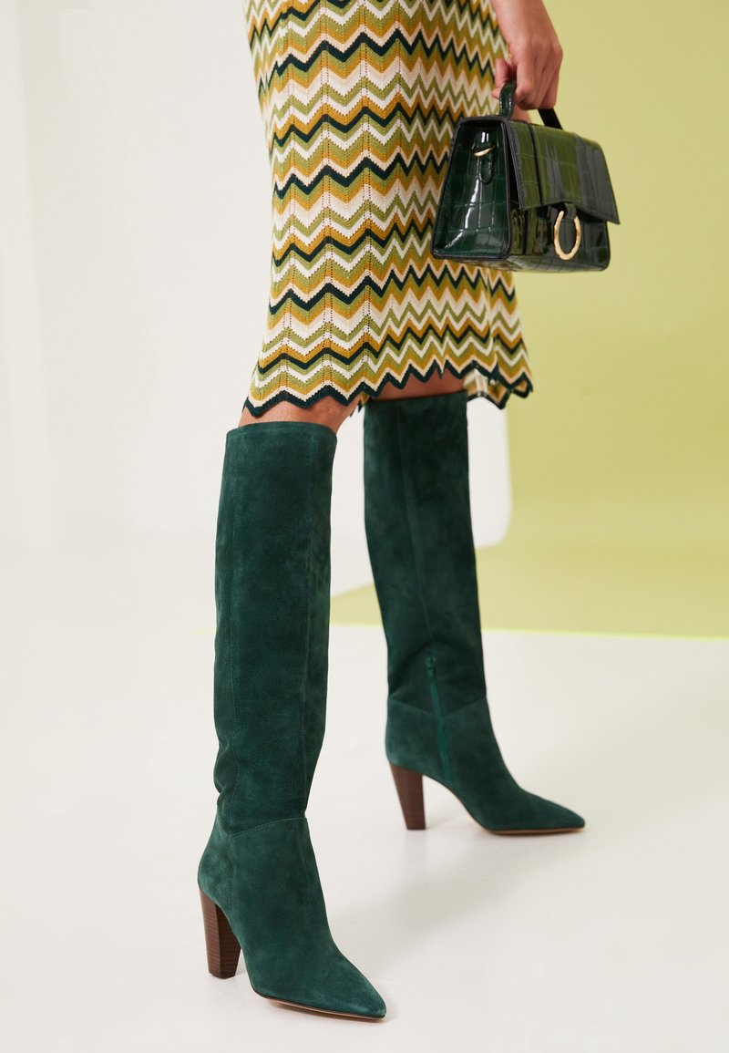 Bottes vertes en suède à talons hauts jusqu'aux jambes, associées à une jupe à motif zigzag et un sac à main vert foncé tenu à la main.