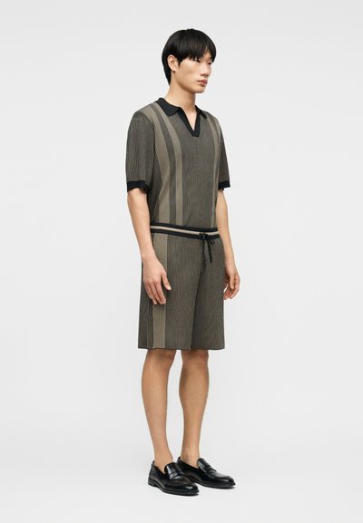 Νέος άνδρας που στέκεται, φοράει ριγέ πλεκτό τοπ με κοντά μανίκια και αντίστοιχα σορτς, με μαύρα μοκασίνια, σε απλό φόντο.