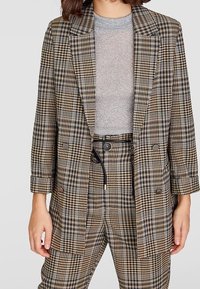 Dvoudílný kostkovaný blejzr v hnědé, béžové a námořnické modři. Podšitý, s klopovým límcem a velkými knoflíky. Nosený přes světle šedý top.