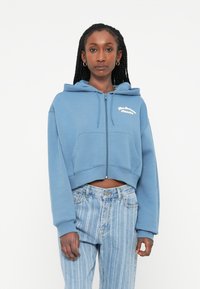 Sweatshirt azul crop com fecho éclair em tecido suave, com um bolso frontal e texto bordado em branco; combinado com jeans listrados claros.
