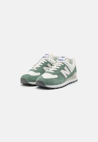 Ett par gröna och off-white New Balance 574 sneakers med ovandel i mocka och mesh, vita snören och svarta gummisulor på vit bakgrund.