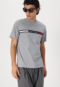 Grå bomulds T-shirt med korte ærmer, prydet med et horisontalt stribet design i rød, hvid og marineblå med teksten "TOMMY HILFIGER".