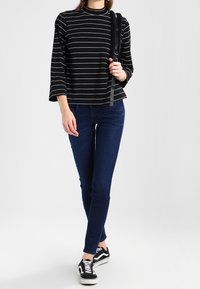 Nainen pukeutuneena musta-valkoraitaisiin pitkänhihaisiin yläosaan, tummansinisii tiukkoihin farkkuihin, musta-valkoisiin tennareihin, kantaa mustaa olkainta olkapäällä.