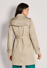 Vrouw met lang donker haar, gekleed in een beige trenchcoat met riem, gezien van de achterkant, tegen een effen lichte achtergrond.