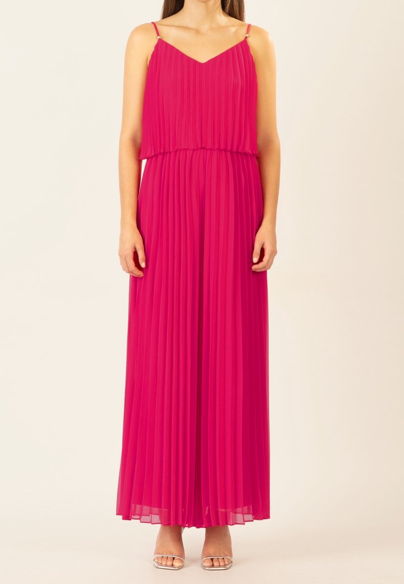 Femme portant une robe maxi rose vif plissée sans manches, avec de fines bretelles et des talons ouverts argentés, debout devant un fond uni.