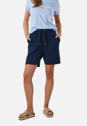 Donna che indossa una t-shirt azzurro chiaro stampata, pantaloncini blu navy con coulisse e tasche, e sandali beige con cinturini incrociati.