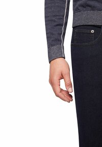 Donkerblauwe trui met contrasterende lichte stiksels, gecombineerd met donker denim broek. Zichtbare hand rustend naast de broekzak.