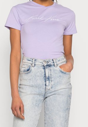 Personne portant un t-shirt Karl Kani violet clair rentré dans un jean taille haute bleu clair effet délavé avec une main dans la poche.