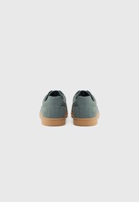 Zapatillas de ante verde con un diseño de corte bajo, suela de goma marrón y acentos traseros minimalistas. Textura suave, forma aerodinámica.
