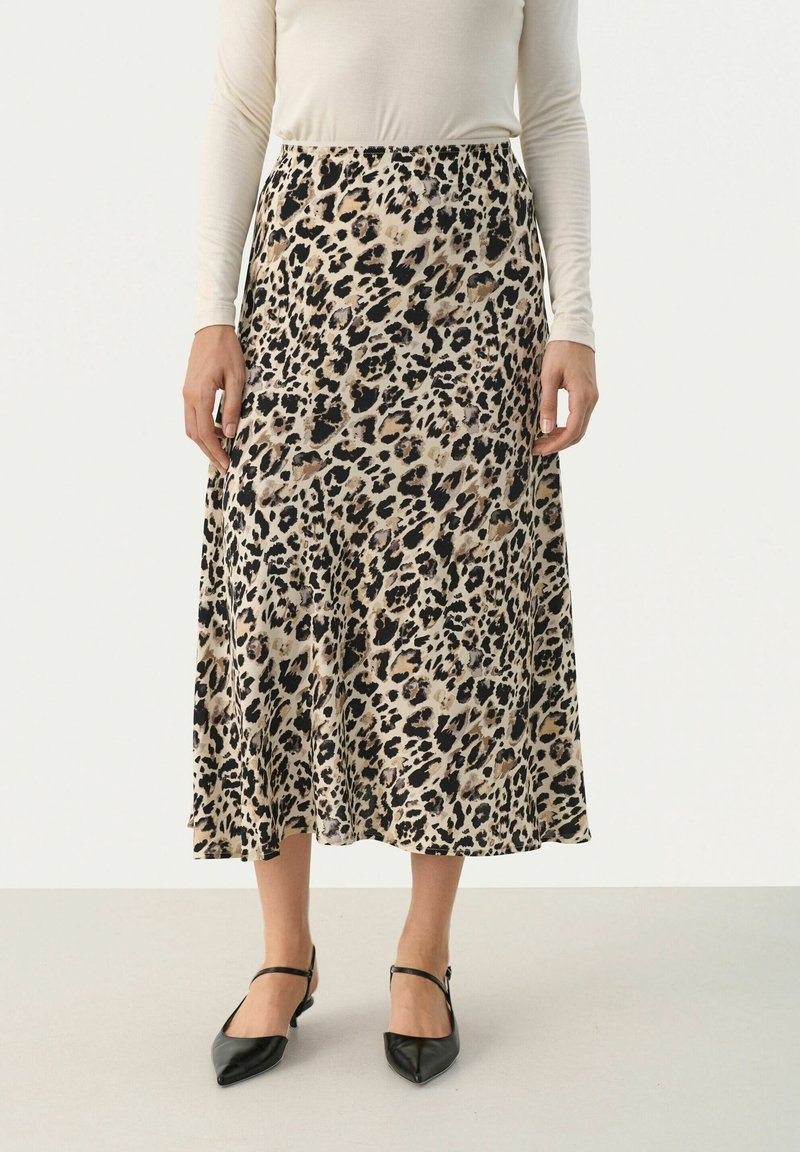 Femme portant une jupe midi beige et noire à imprimé léopard, un haut blanc à manches longues et des chaussures Mary Jane noires à bouts pointus.