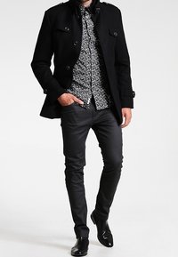 Czarny dwurzędowy płaszcz na wzorzystej czarno-białej koszuli, zestawiony z wąskimi czarnymi spodniami i błyszczącymi czarnymi butami.