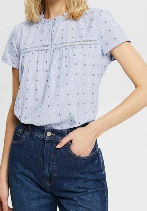 Femme portant un chemisier bleu clair à manches courtes avec petits pois et un jean bleu foncé, une main dans la poche, sur fond blanc.