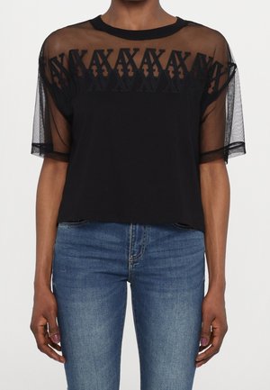 Vrouw draagt een zwart doorschijnend topje met korte mouwen en een geborduurd geometrisch patroon over een effen zwarte stof, gecombineerd met een blauwe spijkerbroek.