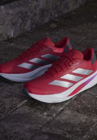 Zapatillas deportivas rojas con parte superior de malla, franjas plateadas y talón gris en contraste. Suela blanca acolchada con un acento rosa en el lateral.