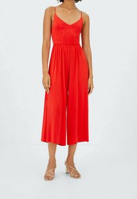 Una tuta rossa con spalline sottili, corpetto aderente e silhouette a gamba larga. Tessuto liscio e design fluido. Abbinata a décolleté dorate con cinturini.