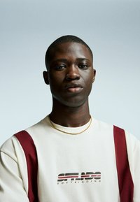 Cream-colored sweatshirt with burgundy shoulder accents. Features printed logo and a gold chain necklace. Plain background.