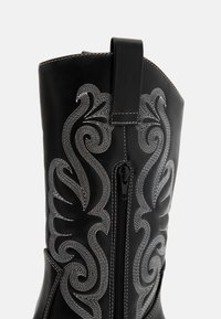 Botte en cuir noir avec des motifs de broderie blanche détaillés et une fermeture éclair latérale. Inclut une languette en haut pour un enfilage facile.