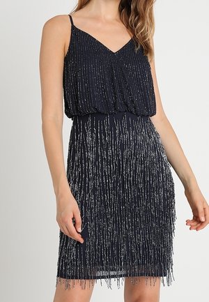 Vrouw in een mouwloze zwarte jurk met dunne bandjes, met verticale zilveren kraalversieringen op de top en rok.