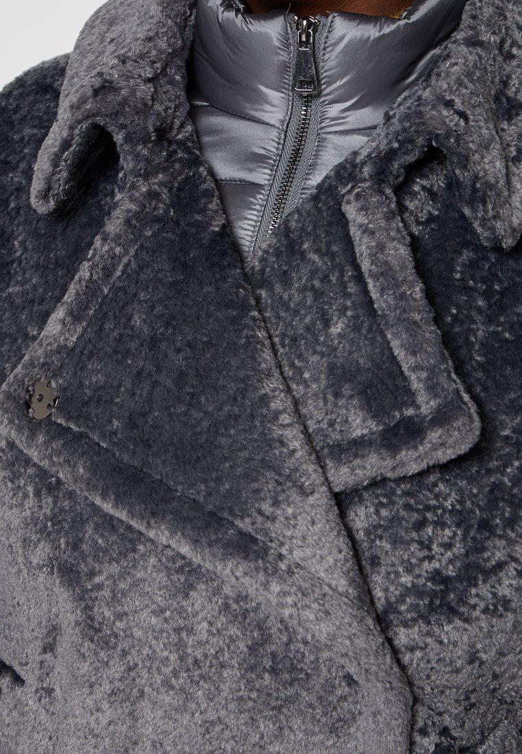 Grau-flauschiger Mantel mit einem strukturierten, weichen Außenmaterial, ausgestattet mit einem grauen Reißverschluss-Innenfutter und einem auffälligen Kragen mit silbernem Knopfdetail.