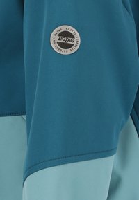 Teal sportjakke med en broderet lyseblå panel og et rundt sølvlogo med "ZIGZAG" tekst på ærmet.
