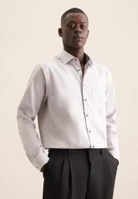 Lichtgrijze lange mouwen shirt met een kraag, voorzien van een contrasterende gestreepte binnenkraag en een linkerknoopzak. Gecombineerd met zwarte broek.