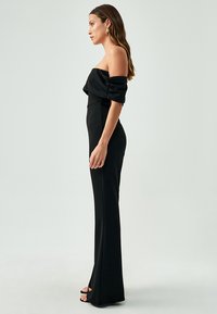 Sort off-shoulder jumpsuit med en tætsiddende silhuet, glat tekstur og vidde ben. Har en subtil samlede detalje nær skuldrene.