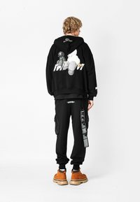 Joven de pie, vistiendo una sudadera negra con un gráfico de dibujos animados en la parte posterior, pantalones cargo negros con etiquetas colgantes y botas marrones, sobre fondo blanco.